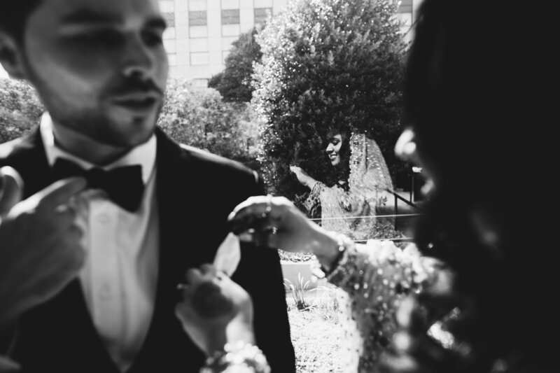 The bride helps the groom with his suit as her smile is reflected in the window glass