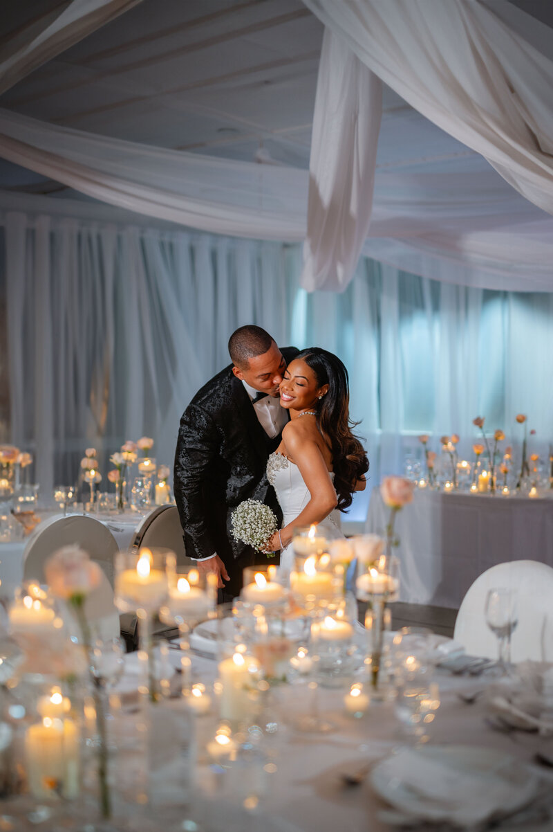 Bride smiles while the groom kisses her cheek in front of a white floral arch.