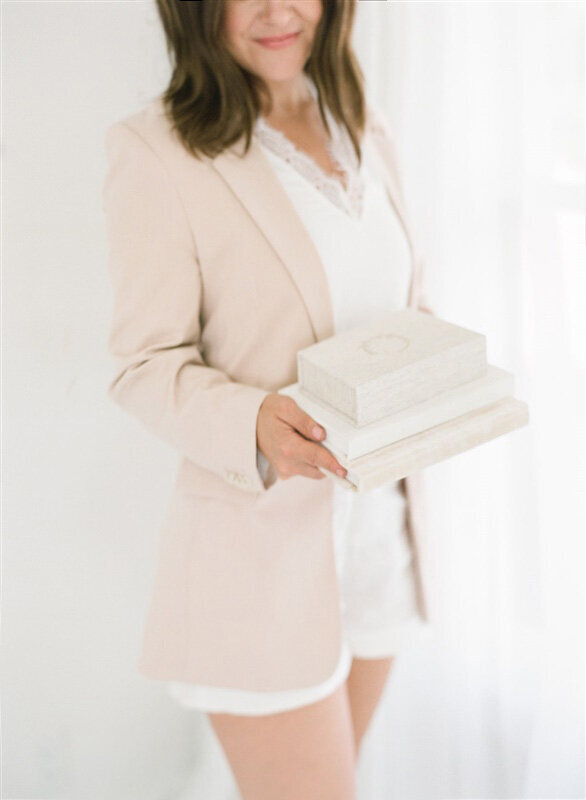 Woman in a soft blazer holding a stack of custom linen heirloom albums — tangible photo keepsakes designed by a Kansas City motherhood photographer.