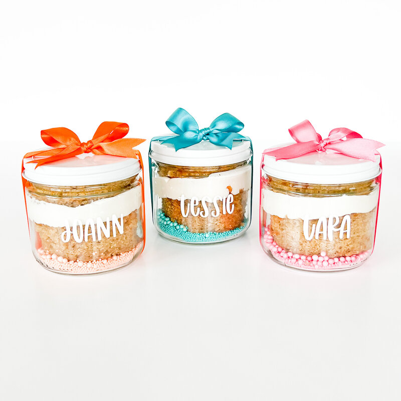 Three cake jars layered with sprinkles, cake and frosting. Jars are tied up with a bow and have a name on the front.