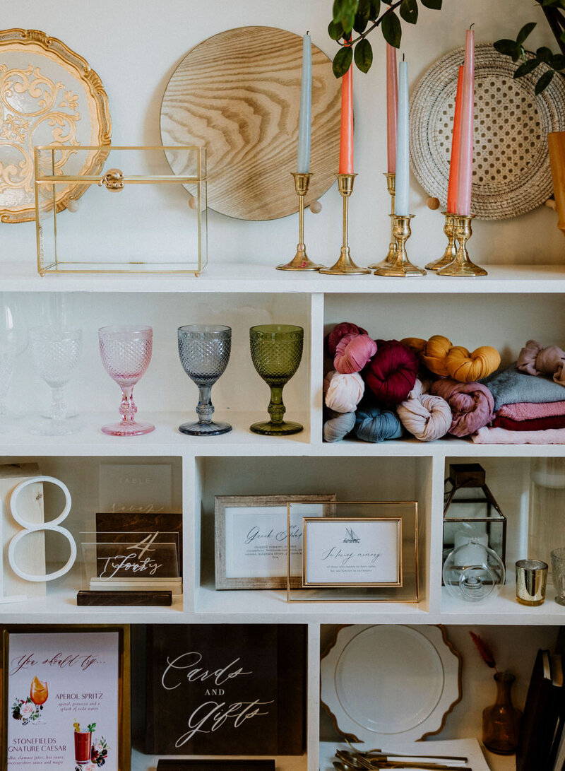 A shelving unit displaying various event rentals like coloured goblets, cheesecloth runners, tapered candles, card boxes and charger plates available to rent are on display at Frid Events' office in Ottawa Ontario.