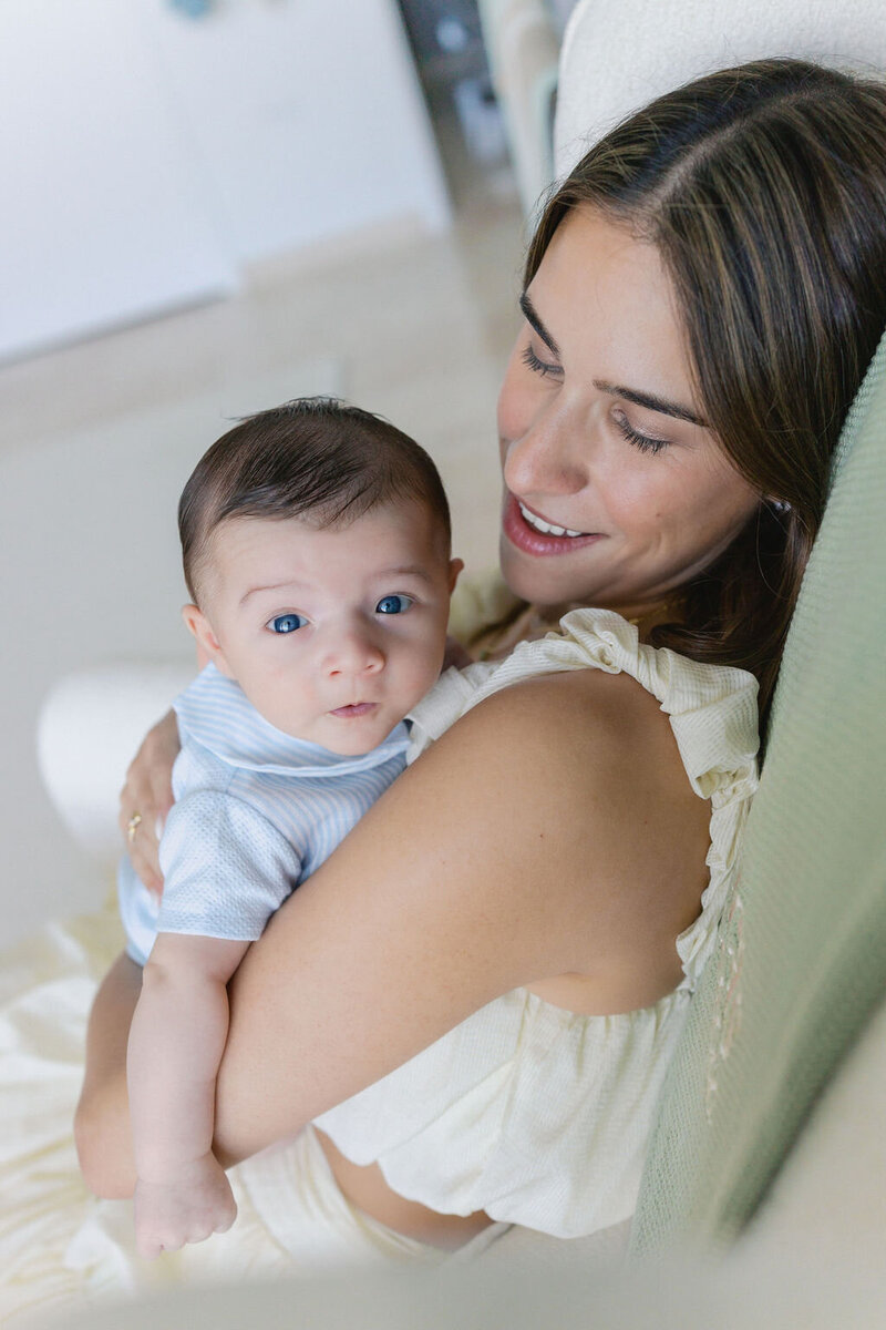Mom sitting in a rocking chair in the nursery and holding her newborn baby who is looking up with his blue eyes in Brickell, FL.