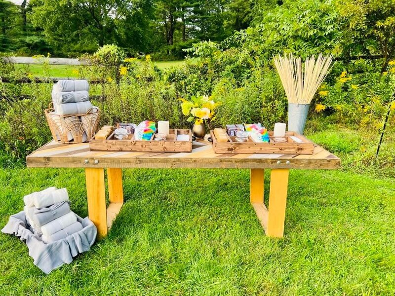 An outdoor event setup featuring a rustic wooden table with blankets, candles, and small gifts arranged on it. A lush green field and trees serve as the backdrop.