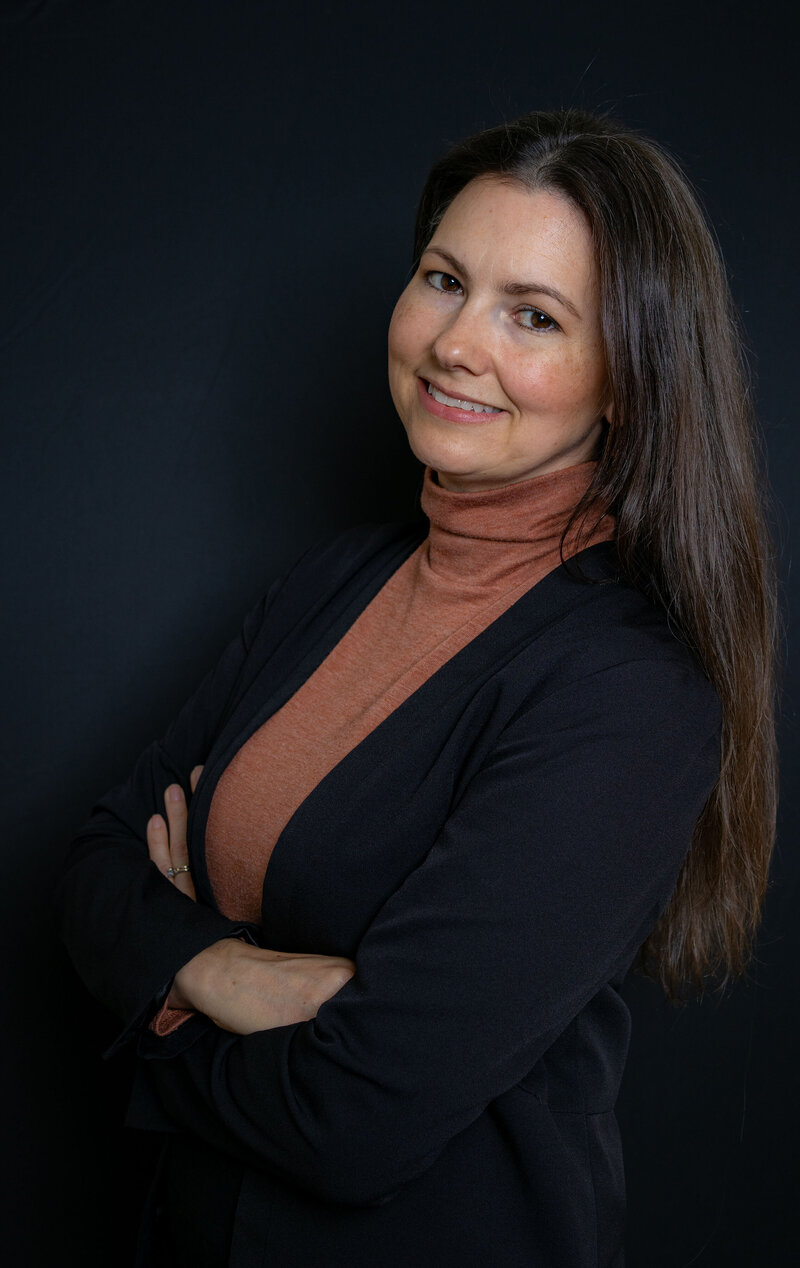 Kellye Mazzoli leaning against a brick column, wearing a navy blazer, rust-colored blouse, and jeans, smiling confidently with a relaxed and approachable pose - City Boss Coaching-Kellye Mazzoli
