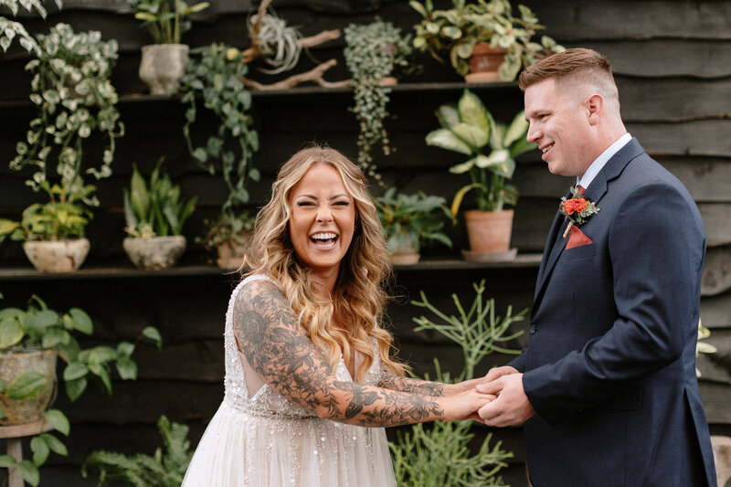 A couple stands together in a greenhouse, surrounded by lush greenery and holding a vibrant bouquet of flowers.