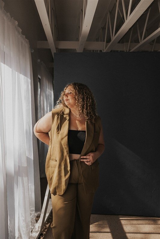 Andrea Cakmar stands in a sunlit room by a window with sheer curtains, dressed in a tan suit and black crop top, gazing thoughtfully out the window.