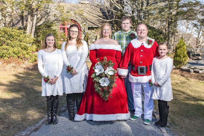 A cheerful outdoor family wedding portrait featuring a bride in a red Christmas dress and groom in a Santa outfit, flanked by children and teens in white dresses and a guest in a green plaid shirt, with rustic buildings and trees in the background.