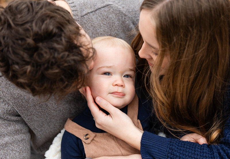 Close-up portrait of baby boy looking at the camera parents kissing him and holding his face during a family session in Farmington, Missouri. 
