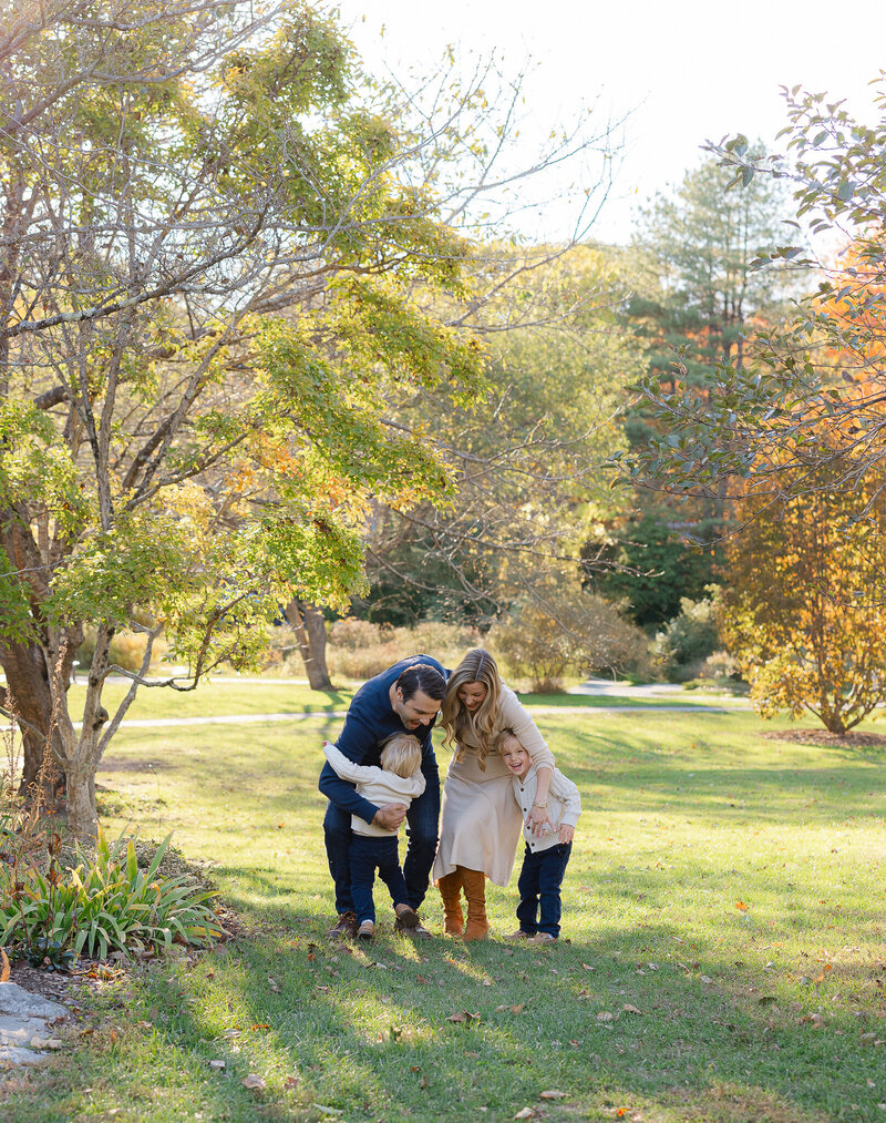 Boston family photographer capturing a joyful outdoor moment of parents hugging their two young children in a sunlit park