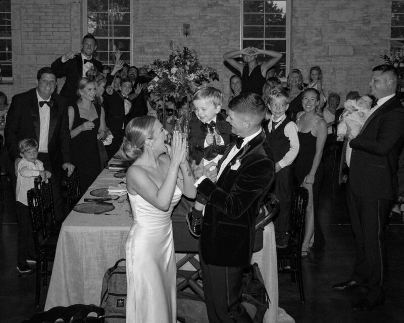 bride and groom together with the guests in the background smiling and laughing while groom holding their son