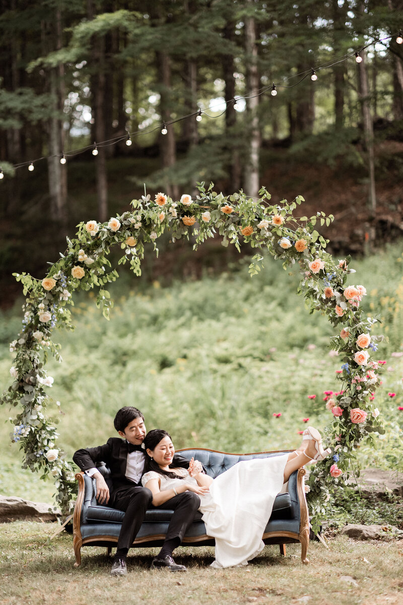 An Asian bride and groom snuggle on a blue velvet couch beside a pond. Behind them is a large circular wedding arbor decorated with pastel wedding flowers.