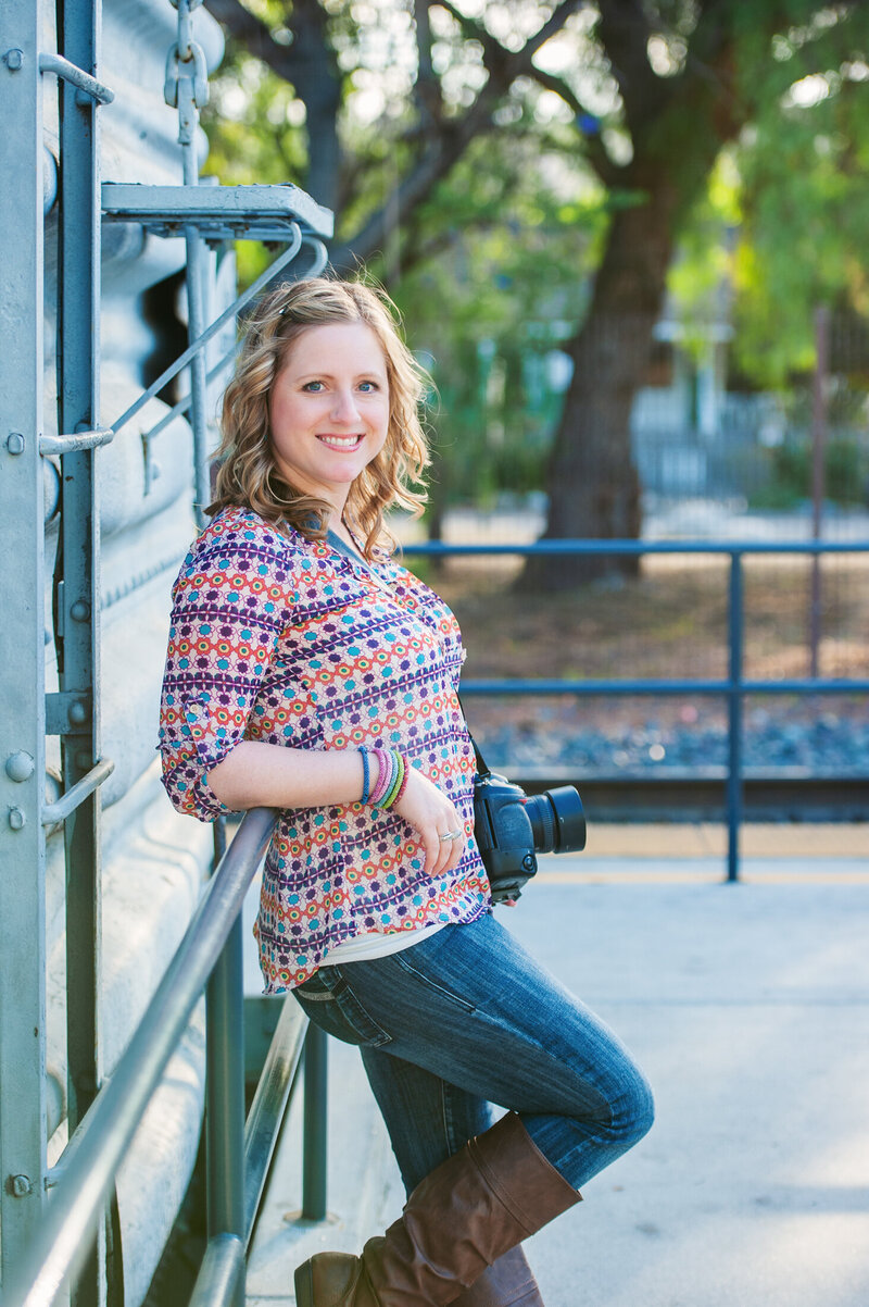 Creative Headshot of Kristin Palkoner, owner of Lens and Prism Personal Branding Photography posed at San Juan Capistrano train station holding Nikon D3s camera.