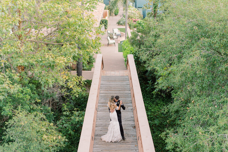 Arial photo of a bride and groom dancing on a wooden bench in a sunny tree filled receptions space.