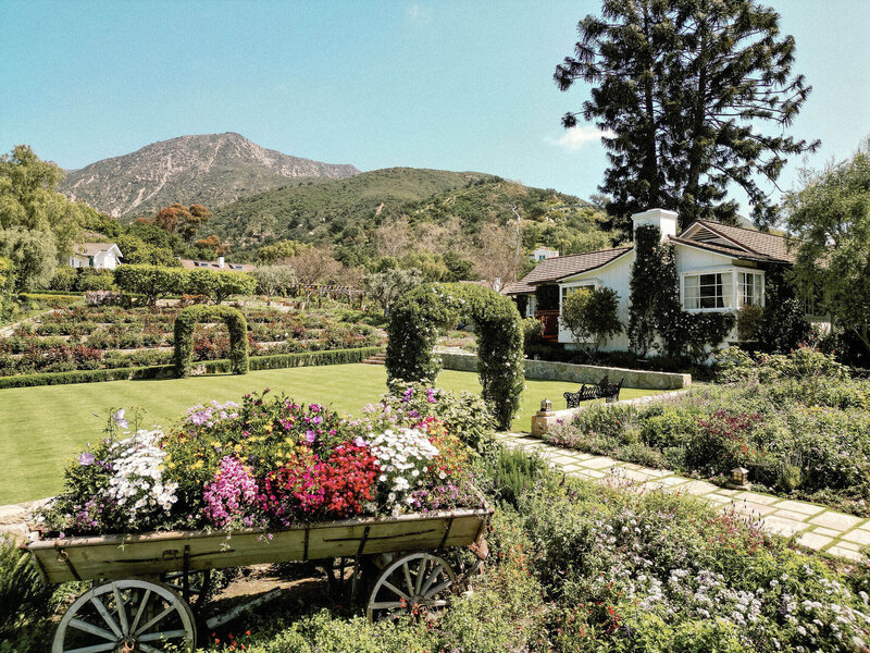 An outdoor view of a Santa Barbara wedding location nestled in the California hills with a large lawn with greenery arches, stone path, gardens, an antique-style wagon filled with flowers, and a white house