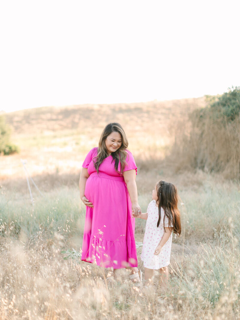 a mom in a bright pink dress holds hands with a young girl in a white dress. They stand in a sunlit field, surrounded by grass and hills, smiling warmly at each other.