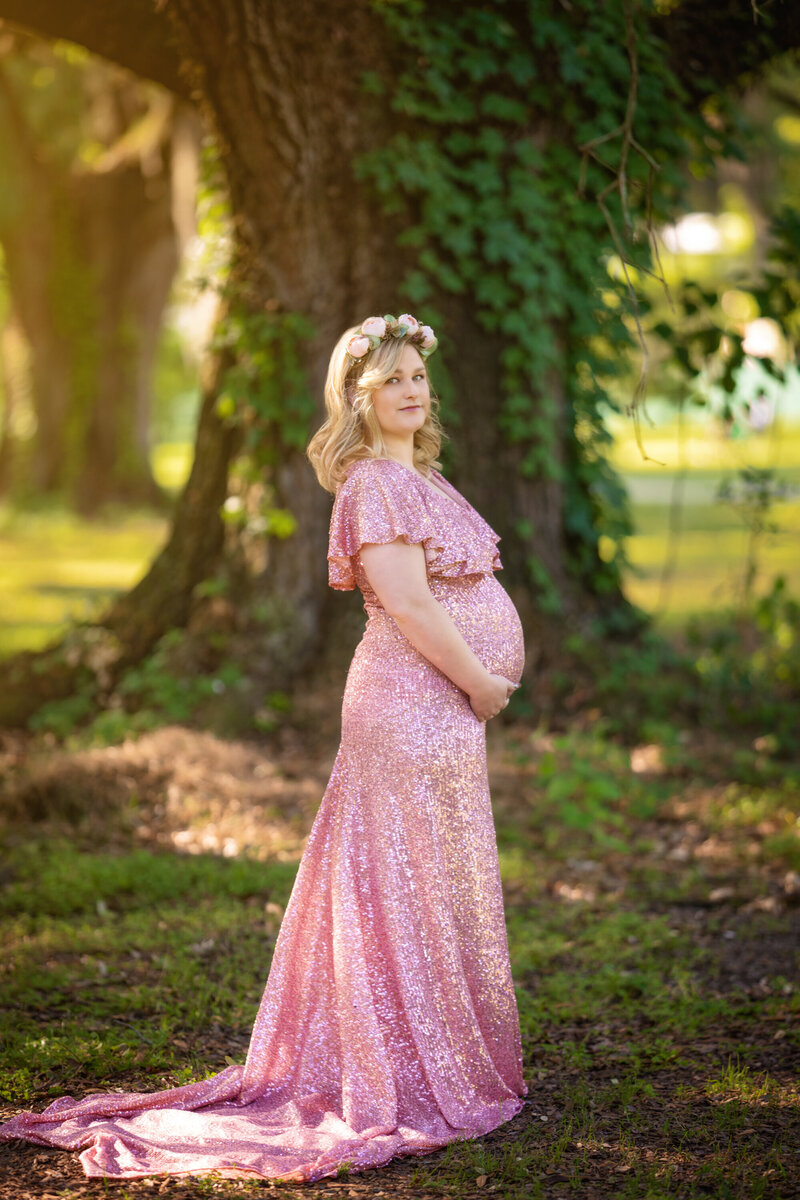 Blond Pregnant woman in a pink sequin dress by Mii-Estillo standing in front of a Live Oak tree in Audubon Park in New Orleans.  She is wearing a flower crown with pink roses and holding her belly.  Her dress has a train behind her.