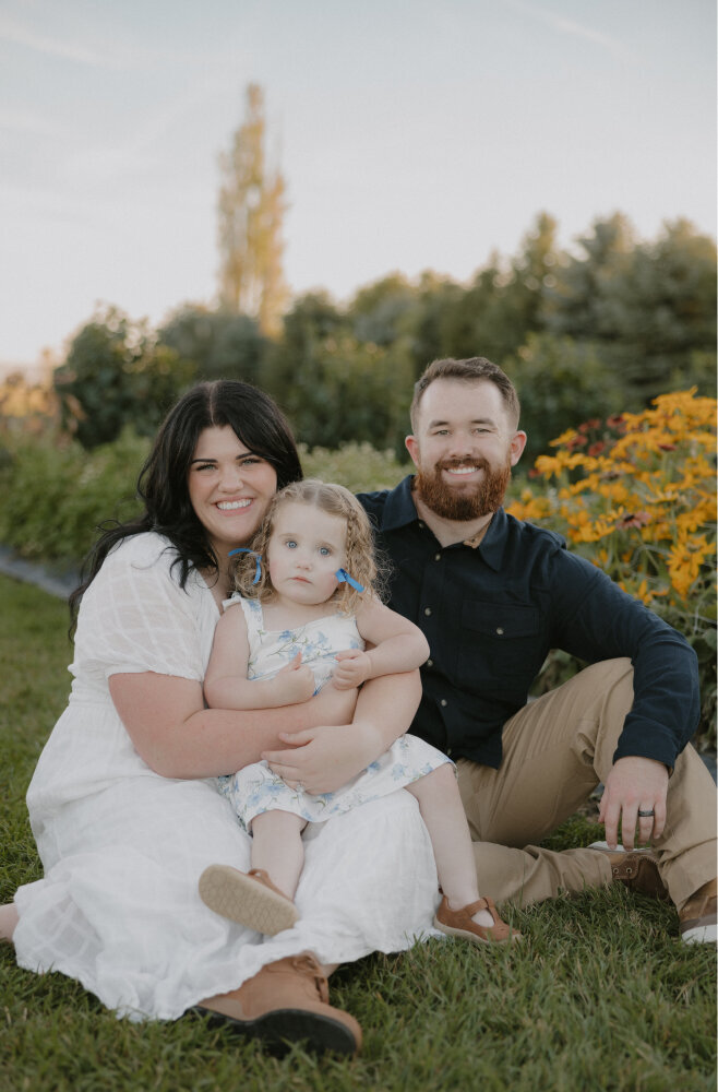 Brigette and her husband are sitting in a grass field, holding their daughter and smiling at the camera.