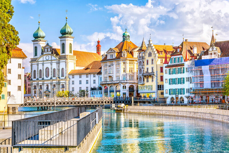 Scenic view of a European town with historical buildings and a river in the foreground. The architecture features ornate facades, a church with green domes, and a bridge crossing the calm water under a partly cloudy sky.