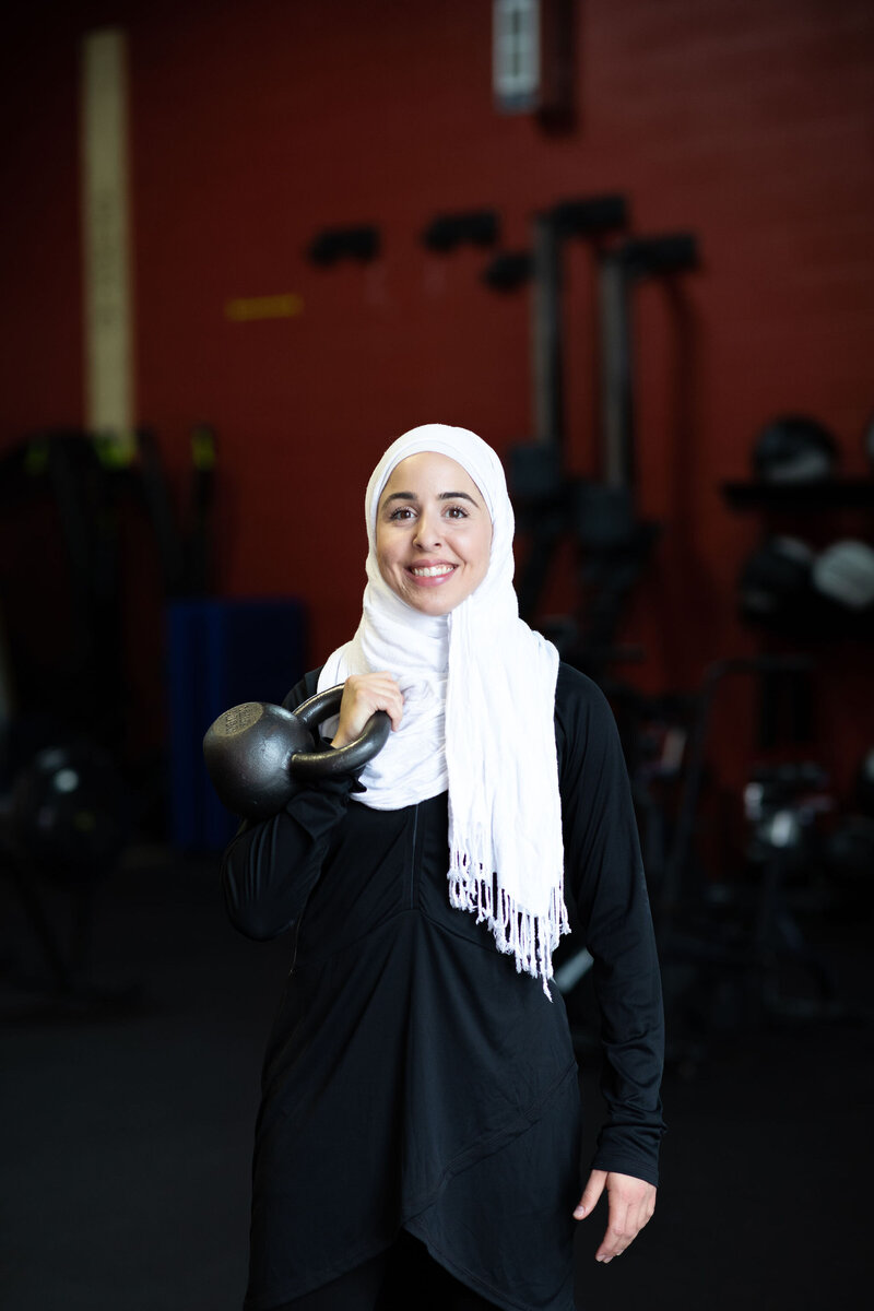 Fitness hijabi coach Hanan posing with a smile while holding a kettle ball inside a gym and wearing black workout gear and a white hijab