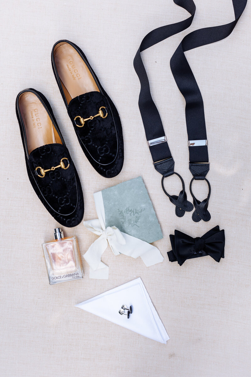 Flat lay of groom's wedding day essentials, featuring black Gucci loafers, elegant suspenders, a classic bow tie, cufflinks, a folded handkerchief, Dolce & Gabbana cologne, and a vow book tied with a white ribbon—all perfectly arranged on a neutral linen background.