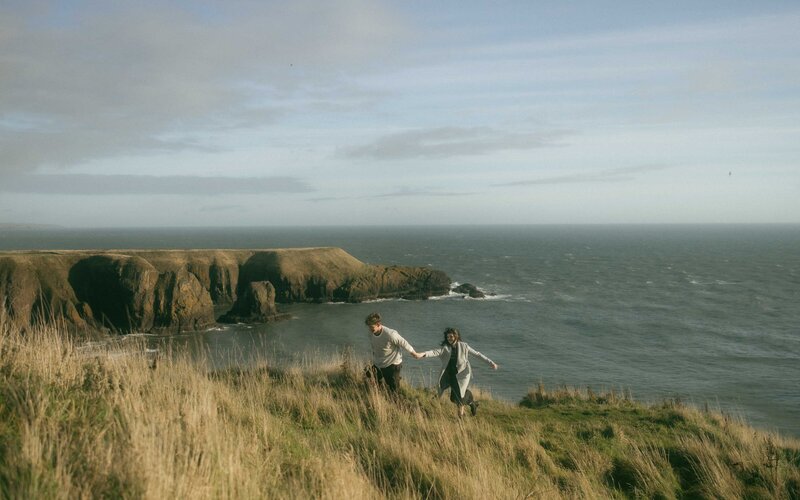 A joyful couple holding hands while running through grassy cliffs by Dunnottar Castle. Captured during a scenic and romantic coastal photoshoot