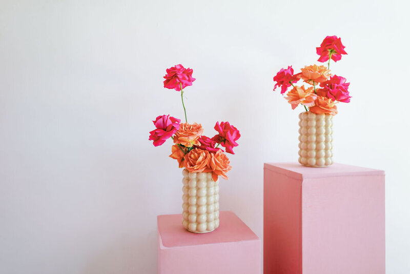 Two white textured vases with pink and orange flowers on top of 2 pink blocks