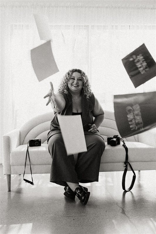 A black-and-white image of Andrea Cakmar sitting on a sofa, playfully tossing photography prints into the air. Two cameras are placed on the sofa beside her.