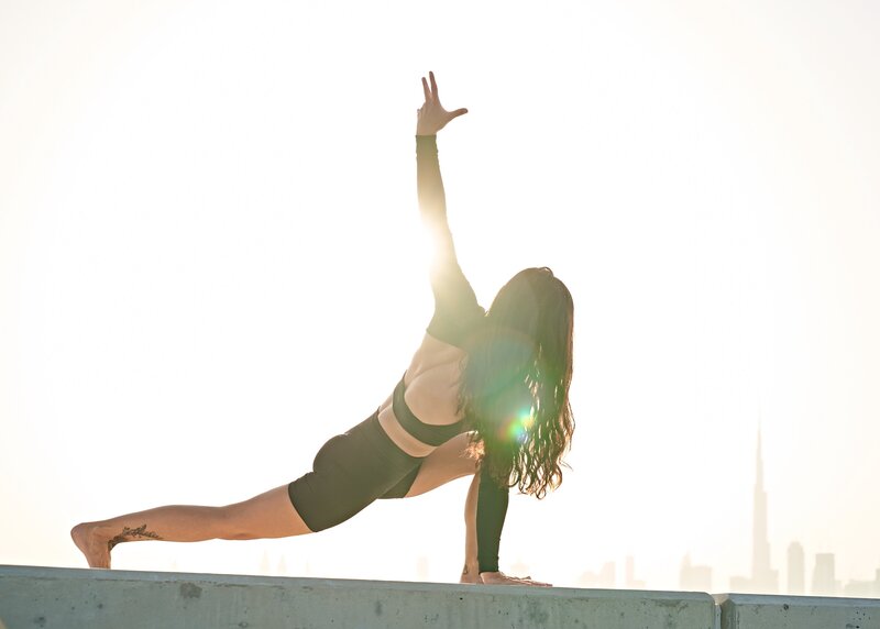 Sarah is performing a strong yoga pose, gracefully balanced on a concrete ledge, with the soft glow of sunrise or sunset framing her silhouette against a city skyline.