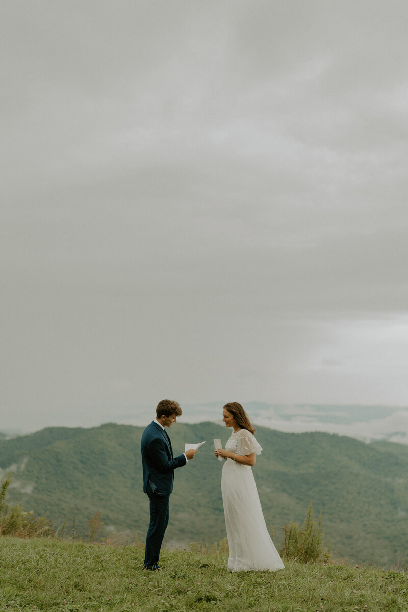 Couple reading vows to each other. The woman is wearing a white dress, the man is wearing a dark blue blazer and white undershirt. In the background there are mountains and clouds.