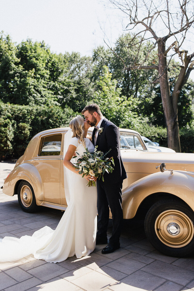Couple de mariés s'embrassent devant une voiture rétro de couleur or. Elle a un magnifique bouquet de fleurs à la main. 