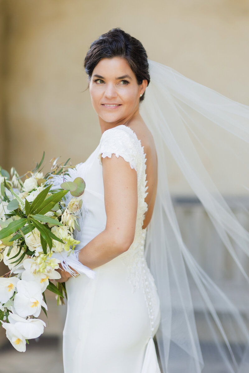 Elegant bride wearing a Pronovias fitted short sleeve dress and a flowing veil is candidly looking over her shoulder