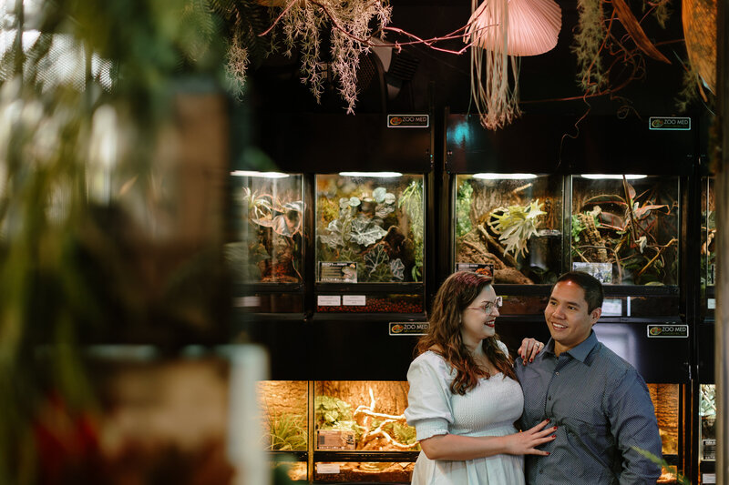 A couple poses together in front of display tanks filled with plants and reptiles, surrounded by lush greenery.