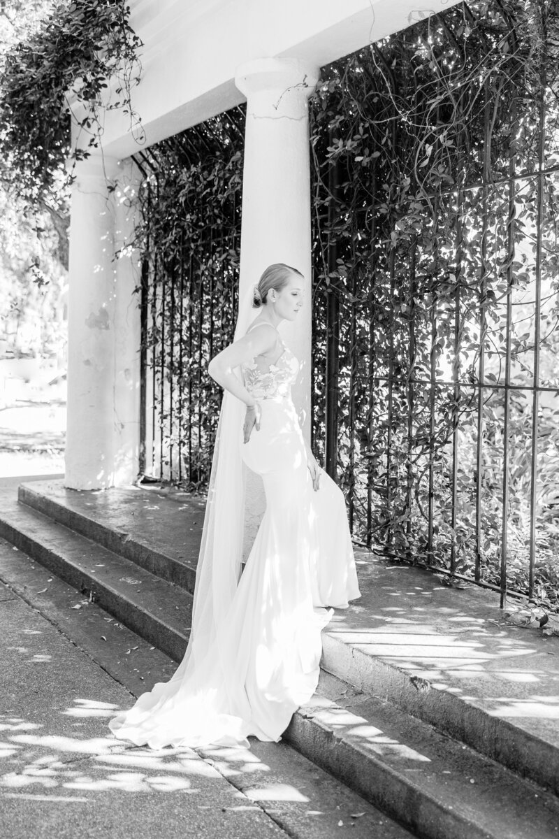 A stunning black-and-white bridal portrait of a bride in an elegant fitted gown with delicate floral details, posing gracefully against a historic column and ivy-covered wrought iron gate in Savannah, GA. Sunlight filters through the leaves, adding a dreamy, romantic touch.