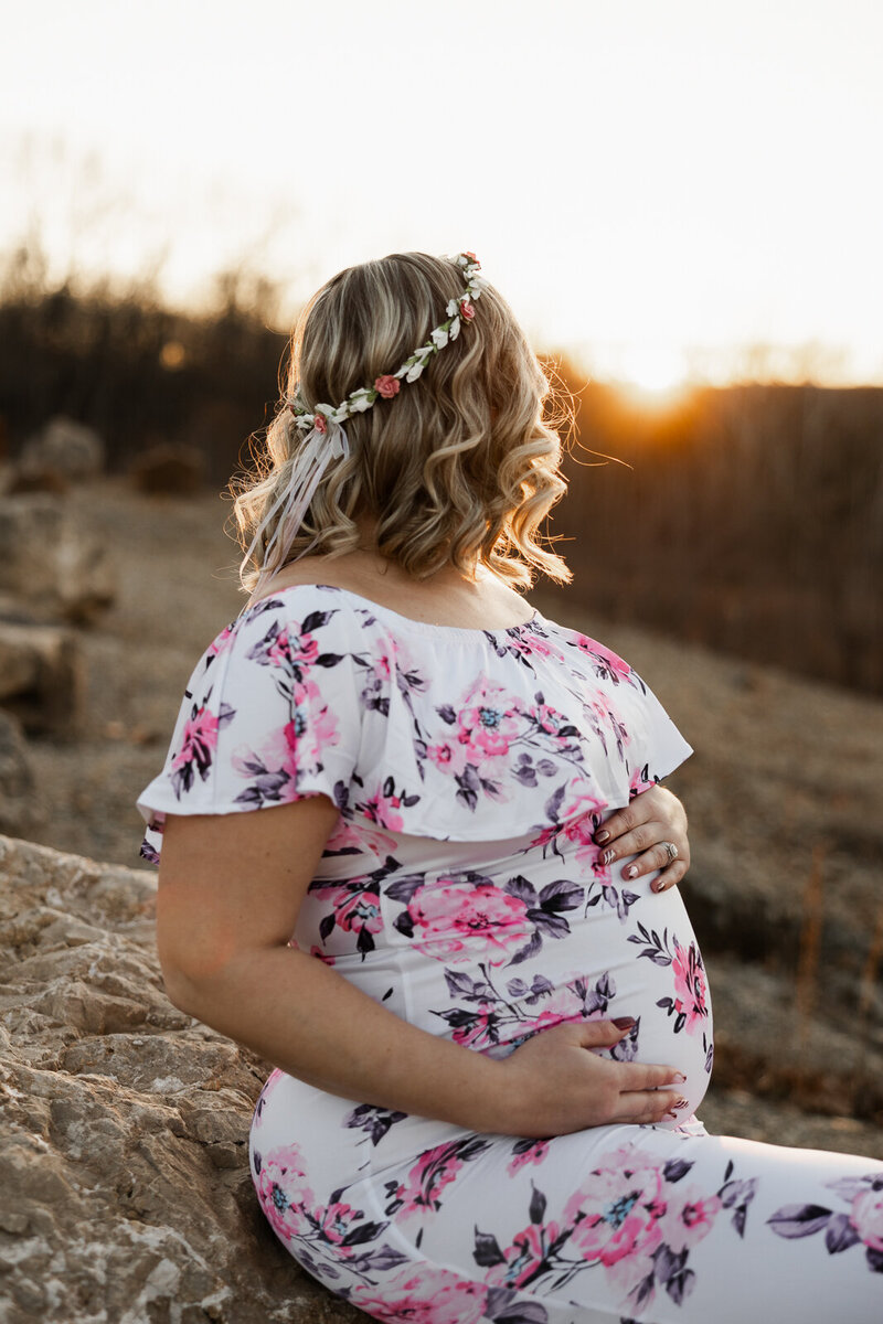 Expecting mother holding her stomach looking at the golden sunset and wearing a floral dress and flowers in her hair taken in Farmington, Missouri. 