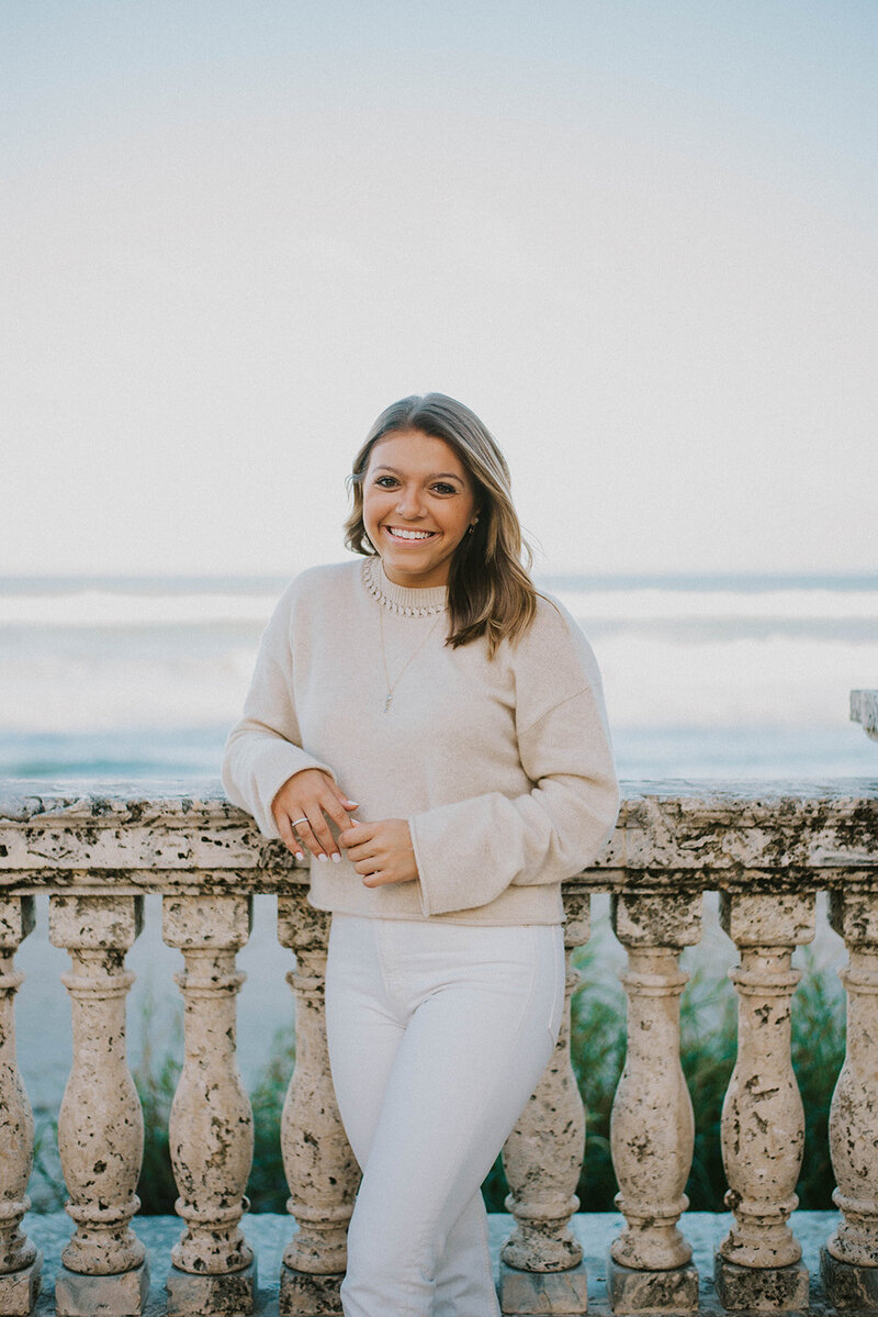Pediatric sleep consultant, Serena Girardi, smiles in front of ocean in tan sweater and white jeans