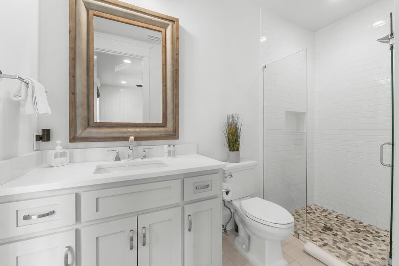 Crisp white bathroom featuring a large framed mirror, walk-in glass shower with pebble tile, and single vanity at Carillon Beach rental.