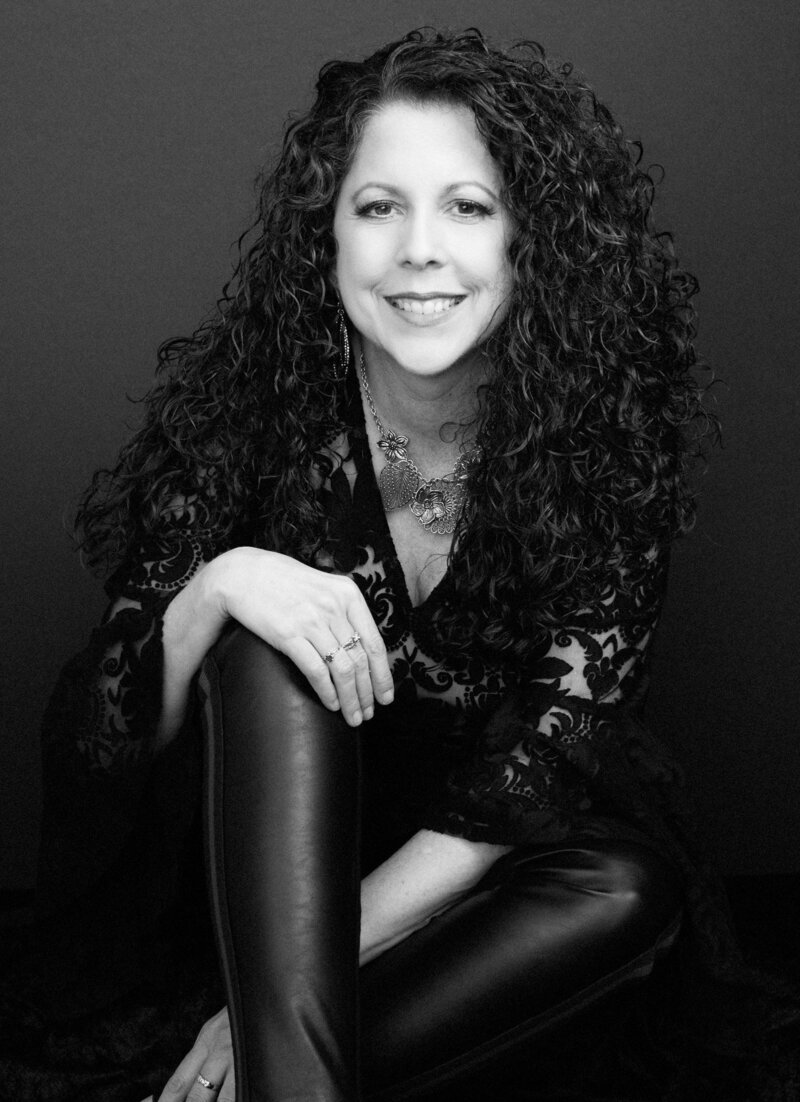 Black and white photo of a smiling woman with long, curly hair, wearing a patterned top, and holding the back of a chair with one hand.