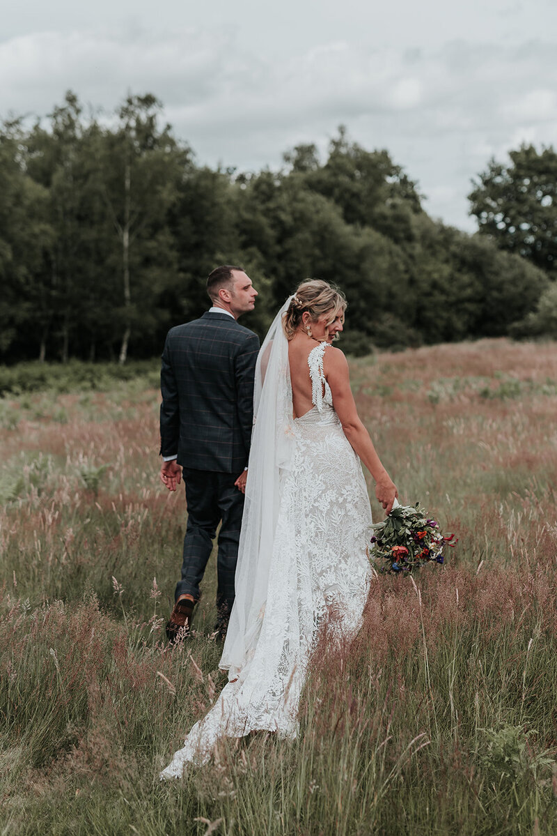 Couple walk through long grass field, brides dress flowing in the grass at Two Woods Estate, Pulborough