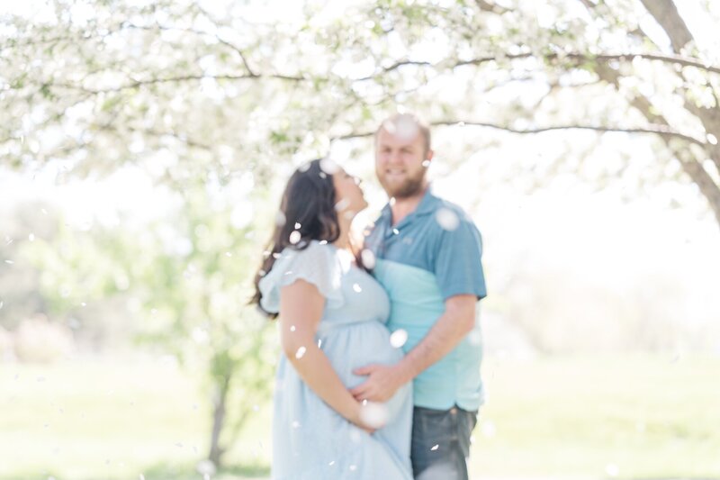 a man smiling while holding his pregnant wife's belly as they stand under a blossoming tree