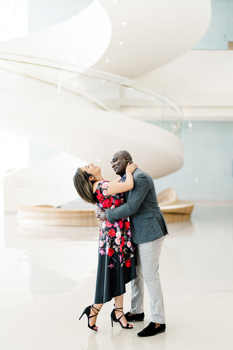 Wedding photographer in Dallas gets a photo of a couple indoors with a large staircase behind them, the girl is laughing and the guy is smiling at her while they hug. She is in a bright floral dress and and he is wearing a gray blazer.