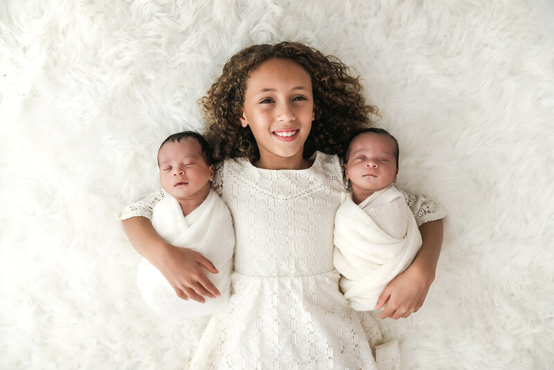A smiling girl with curly hair lies on her back on a fluffy surface, holding two swaddled newborn babies, one on each side. All three are dressed in white and appear content and peaceful.