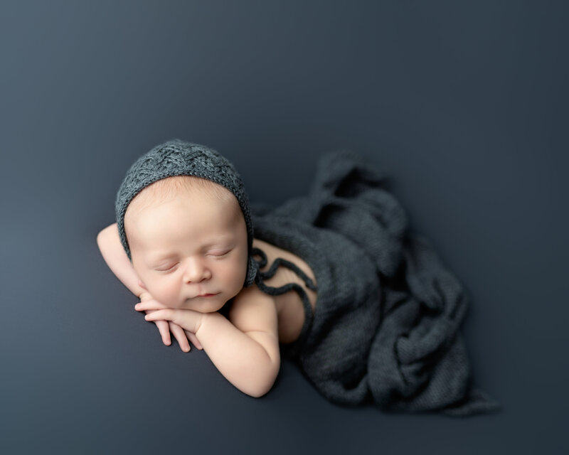 Classic newborn pose with textured knit bonnet on rich grey – softly lit and timeless.

