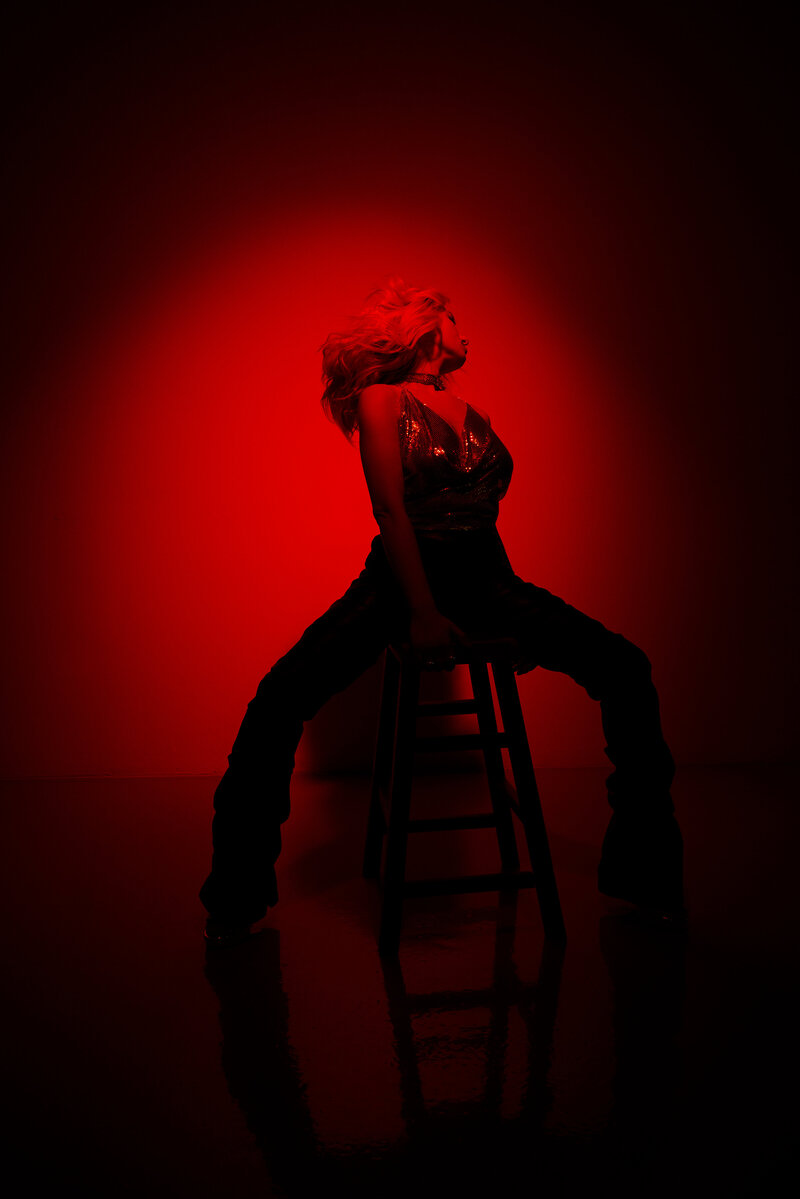 Red light image of tina win sitting on a stool in a photography studio.