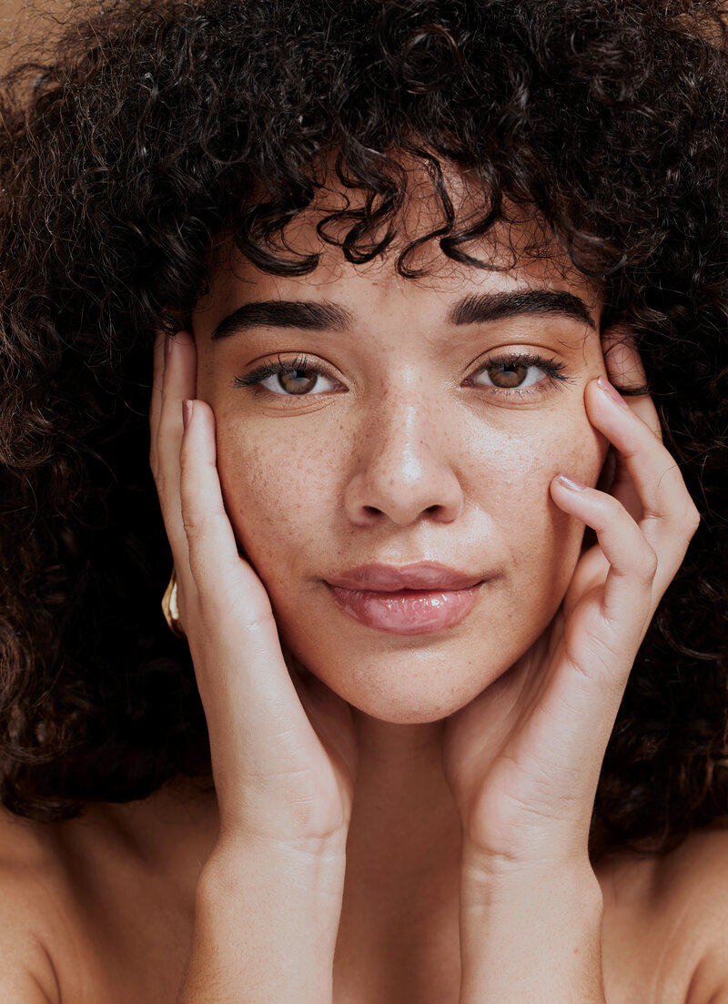 Woman with curly hair and freckles holding her face
