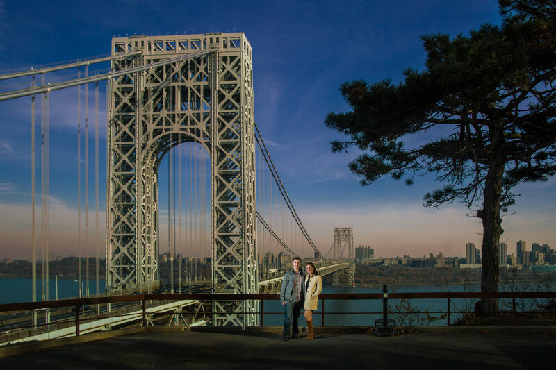 Engaged couple standing near a bridge overlooking the New York City skyline, showcasing their love at this destination engagement session.