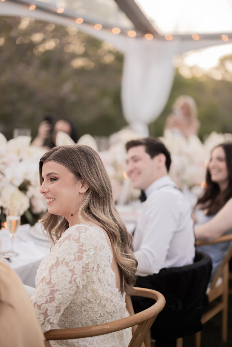 Bride smiles during heartfelt toasts at an outdoor wedding reception under a clear-top tent in the Hamptons, surrounded by floral centerpieces and guests.