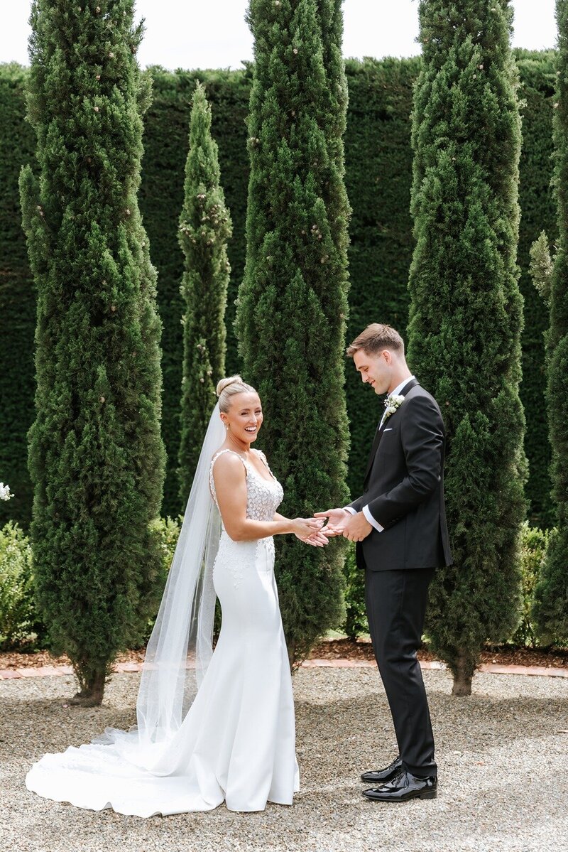 A bride with a long veil is holding hands with her groom in front of hedge trees.