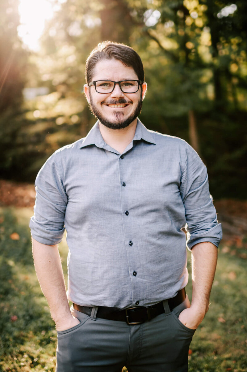 A dark hair man stands in a grassy field with arms crossed wearing a tan long sleeve henley