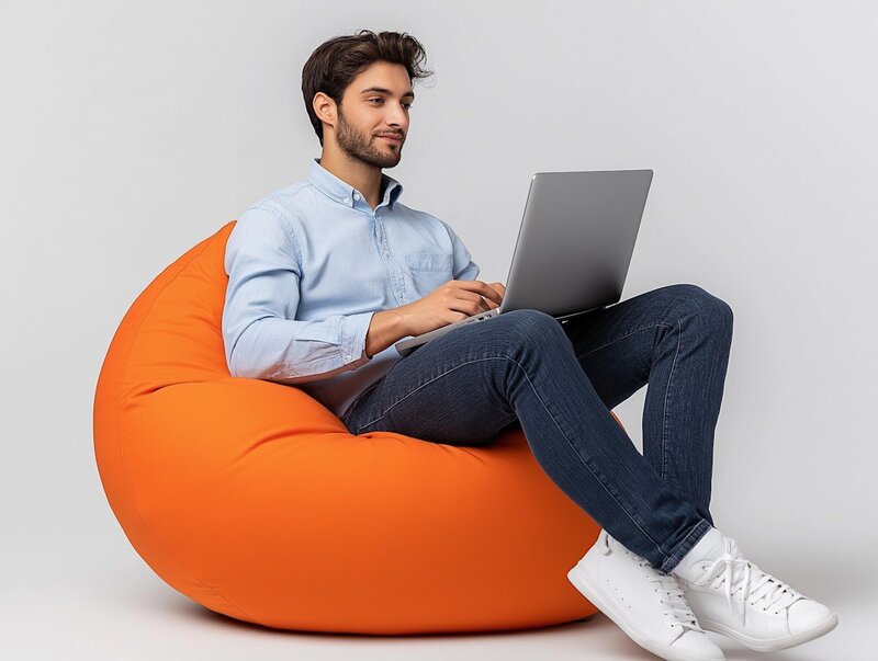 muž sedí na oranžovém sedacím vaku, na klíně má laptop, dívá se do něj a usmívá se