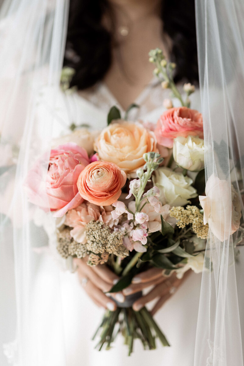 A bride in a veil holds a hand-tied wedding bouquet of pink peach and yellow garden roses and ranunculus
